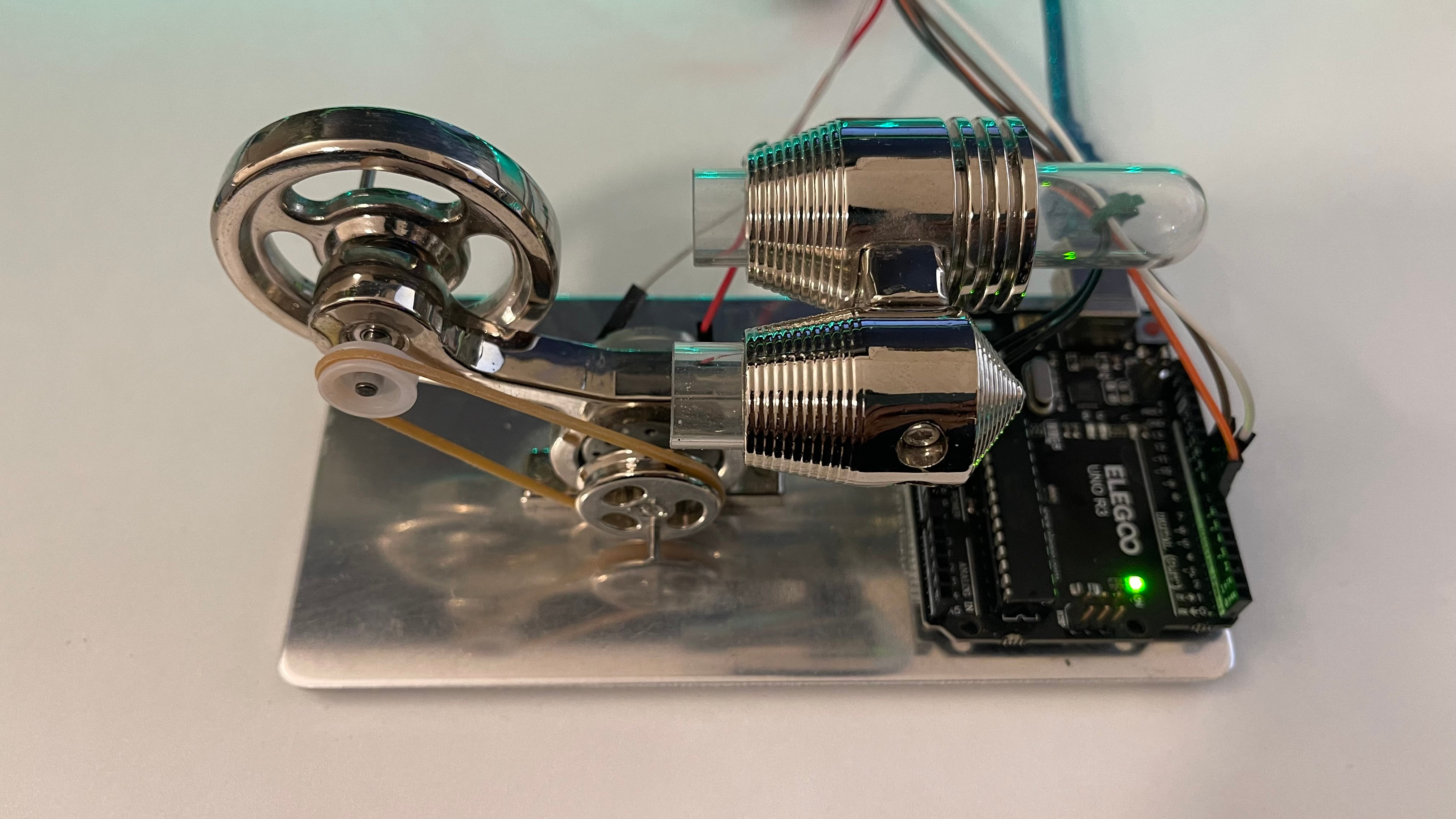 A Stirling-engine chassis with flywheel and drive belt, repurposed as an Arduino-controlled desk toy next to an Elegoo Uno R3
